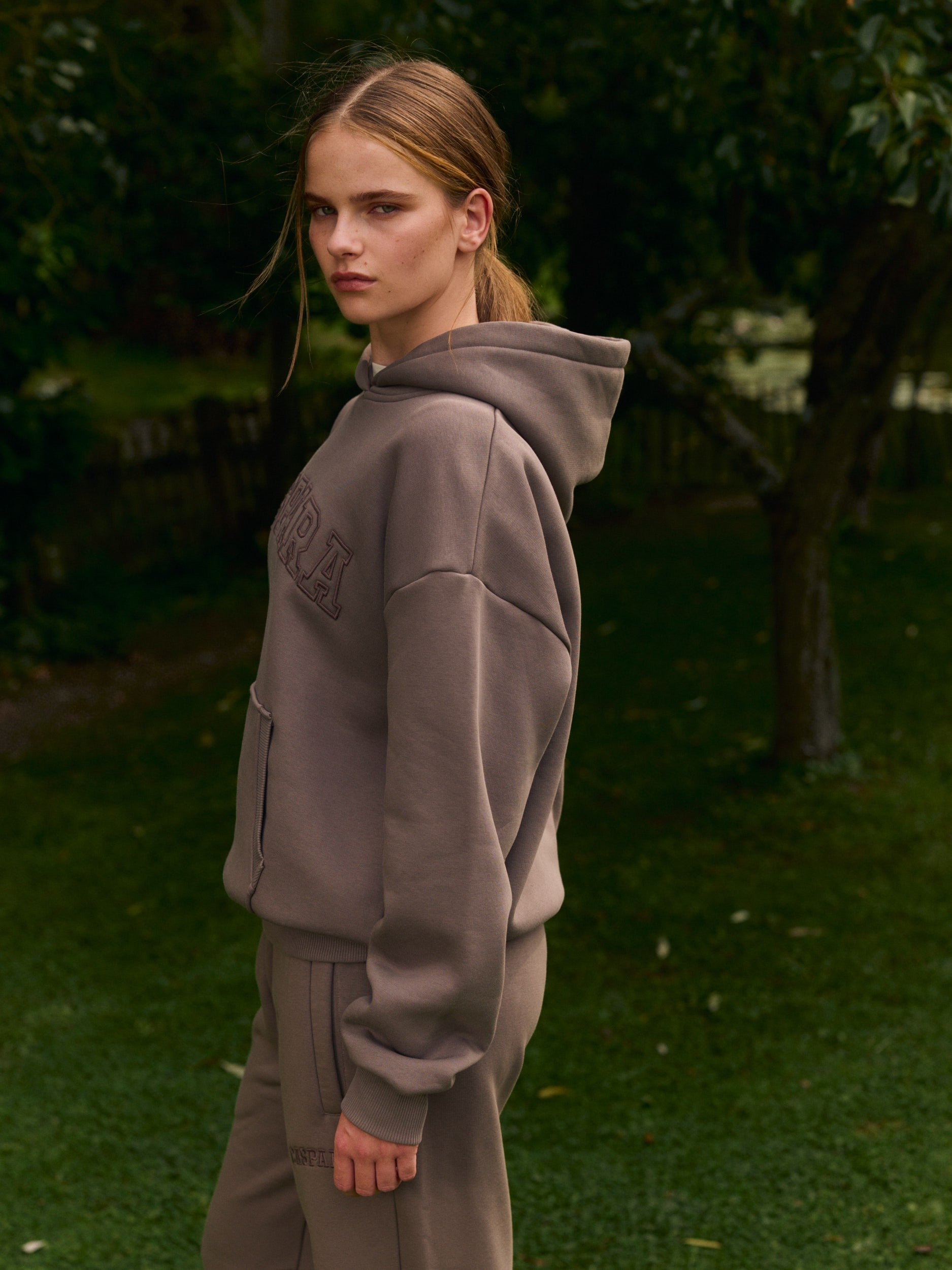 Woman wearing a walnut brown oversized hoodie standing outdoors in a garden.